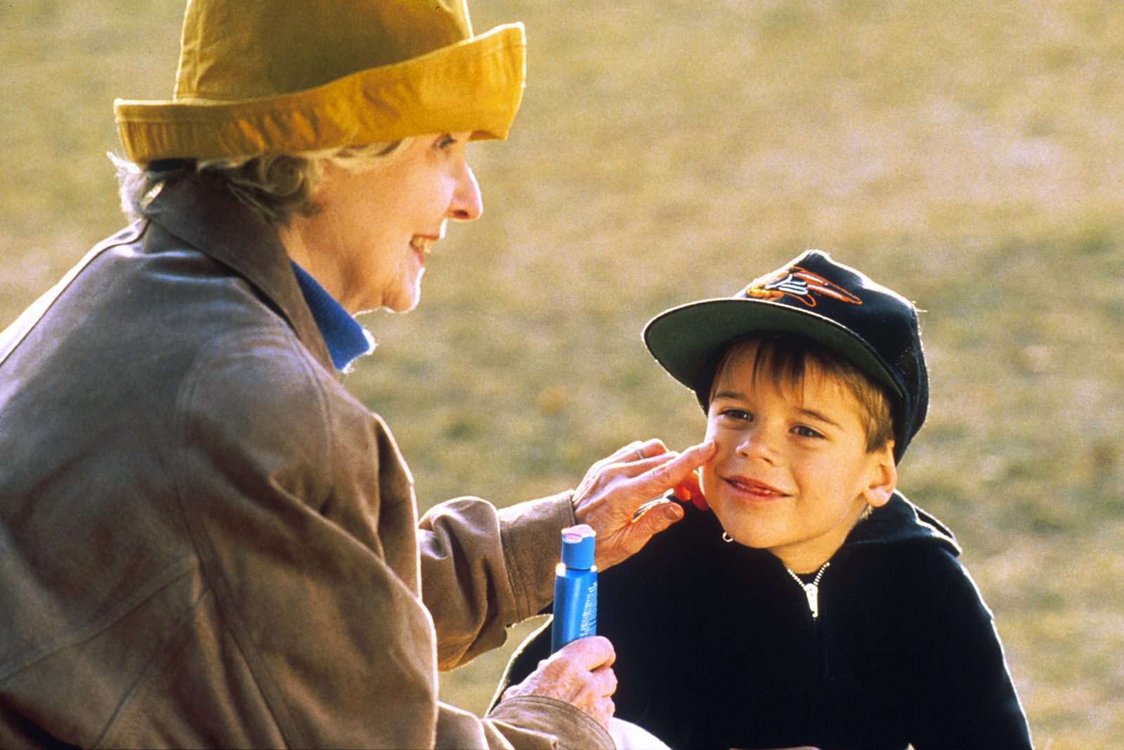 An older woman applies sunscreen from a blue tube to a smiling child's cheek outdoors