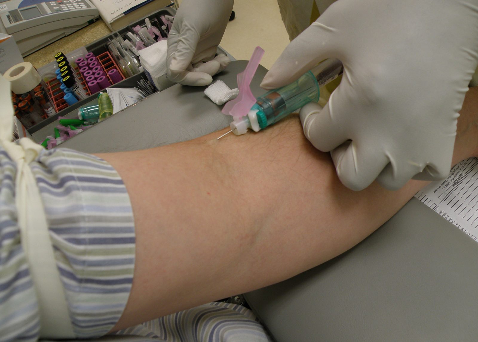 A gloved clinician draws blood from a patient's arm using a vacutainer kit, with sample tubes visible on the counter