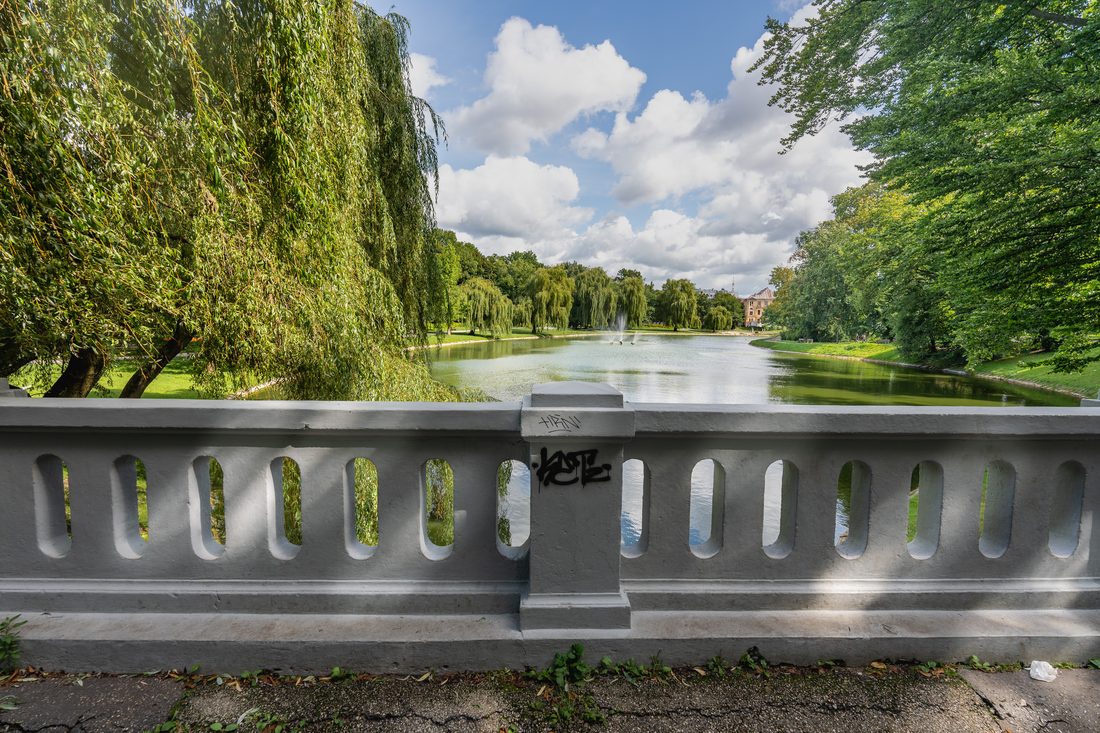 A lush city park with willow trees, a pond, and a fountain under a partly cloudy sky