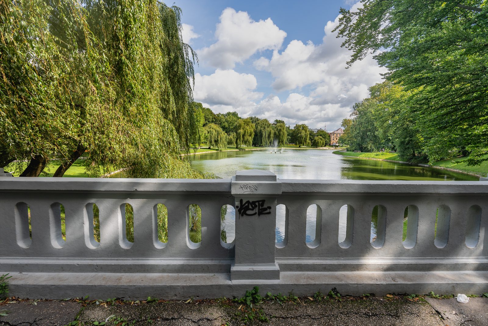 A lush city park with willow trees, a pond, and a fountain under a partly cloudy sky