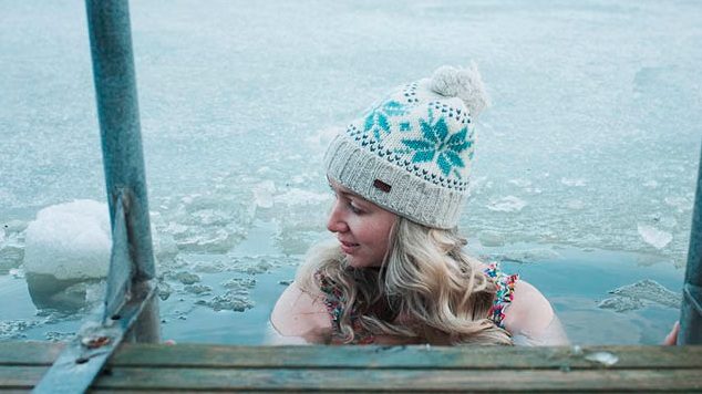 A woman in a winter hat is chest-deep in icy open water dotted with ice chunks, smiling, beside a wooden entry ladder