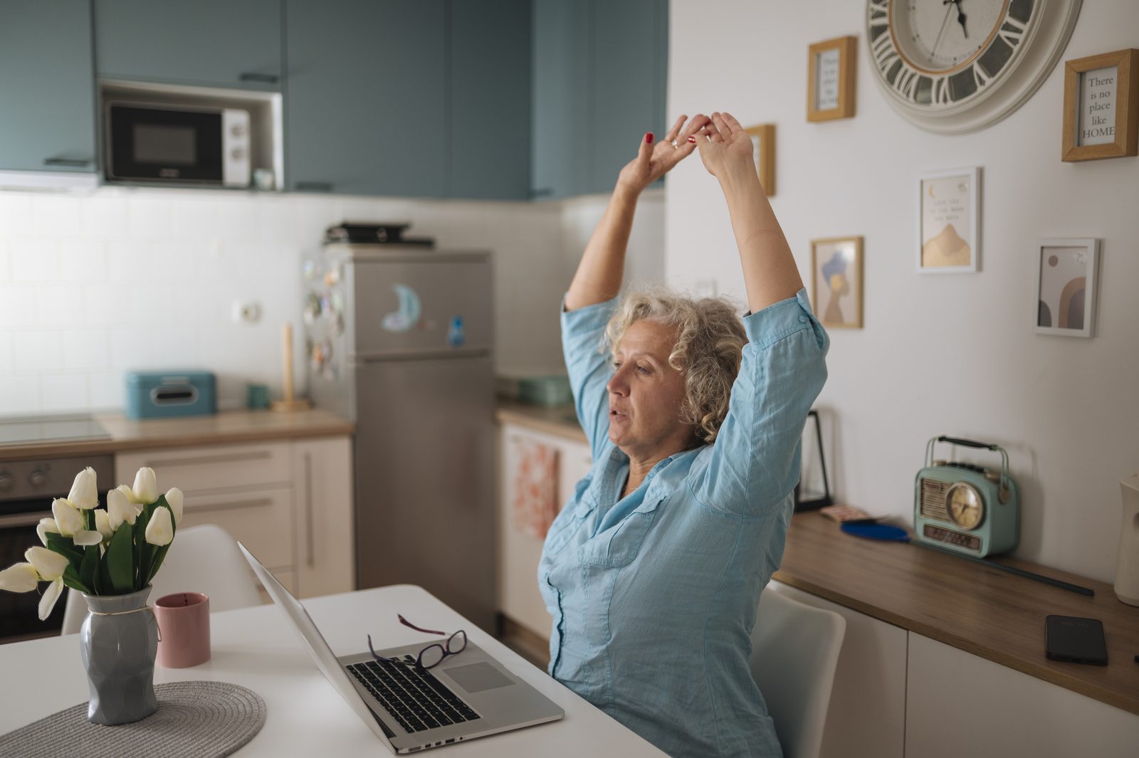 A woman stretches her arms overhead while sitting at a kitchen table desk with her laptop open