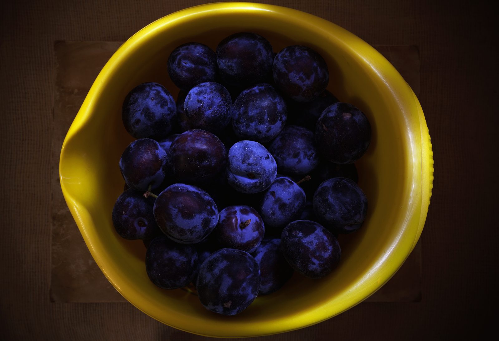 Close overhead view of dark, fresh plums piled in a yellow dish, dramatically side lit