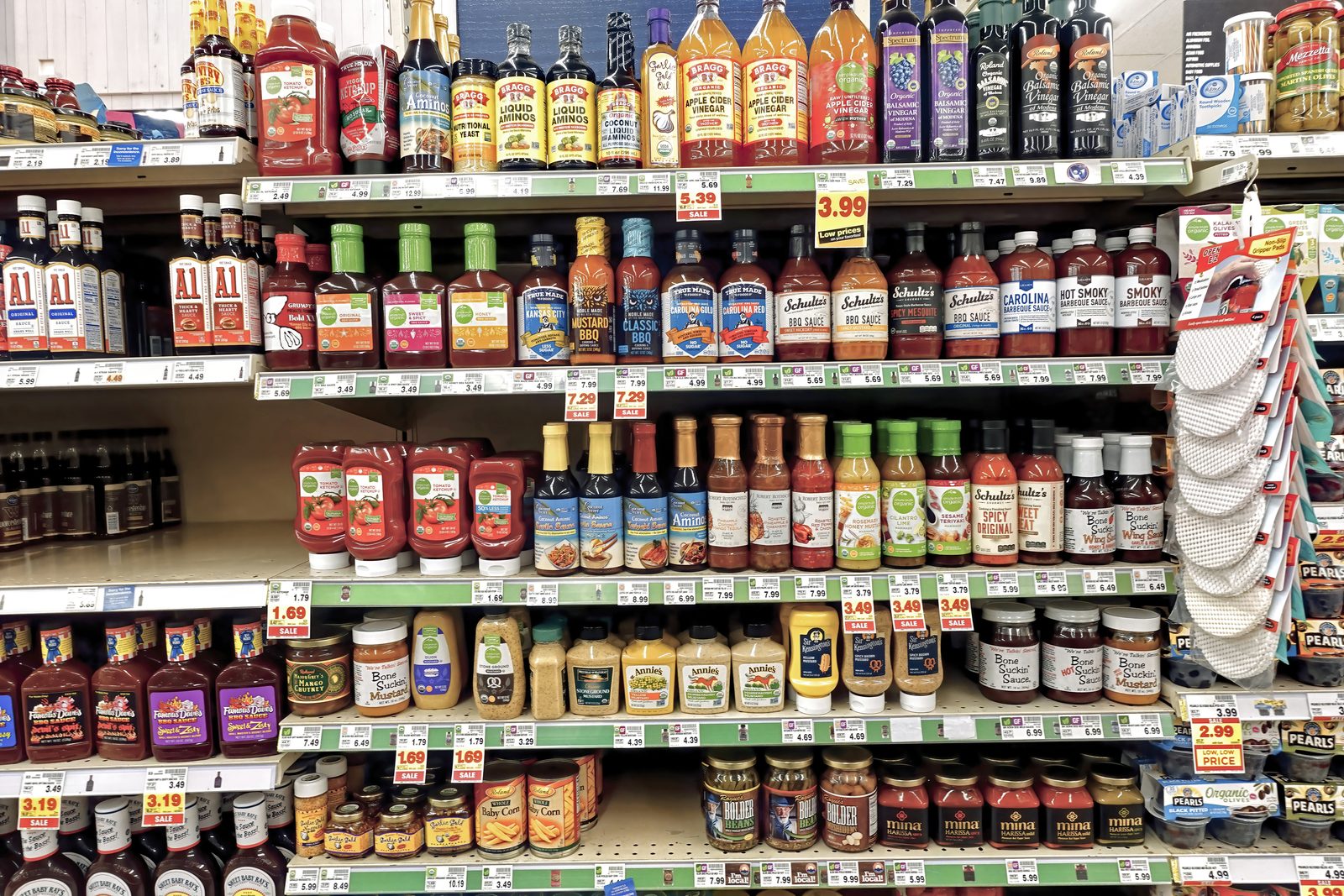 A wide grocery store shelf densely stocked with dozens of different bottled condiments and sauces, ketchups, barbecue sauces and mustards from many different brands