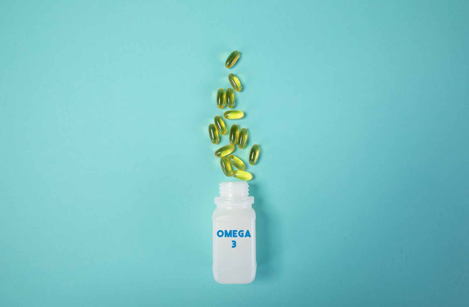 Golden omega 3 fish oil softgel capsules spilling upward out of an open white bottle labeled OMEGA 3, against a teal background