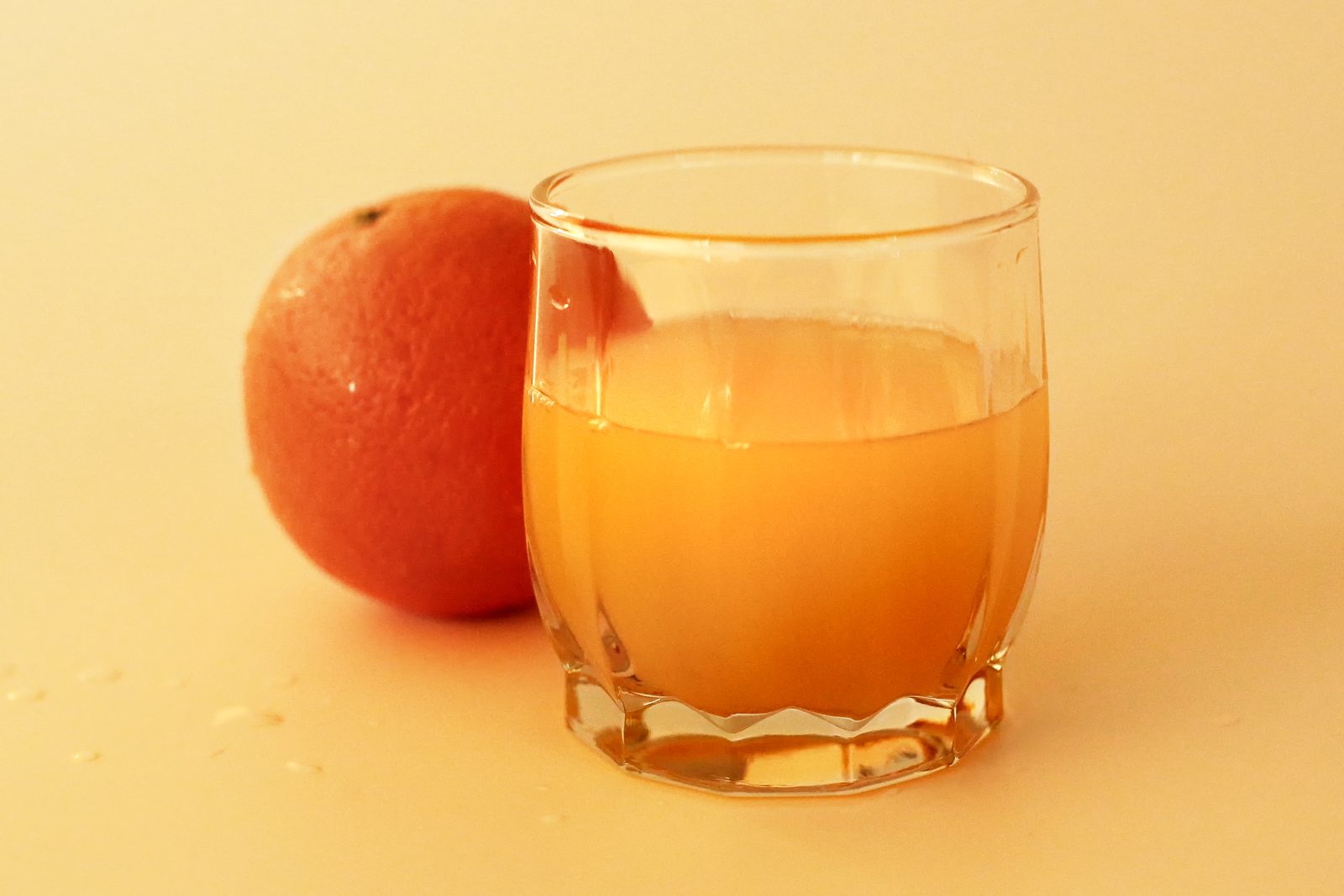 A short glass of orange juice beside a whole orange on a pale warm background