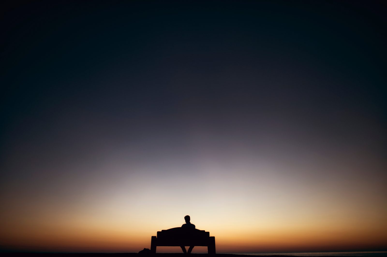 A person sits alone on a bench, silhouetted against a dawn or dusk sky over water