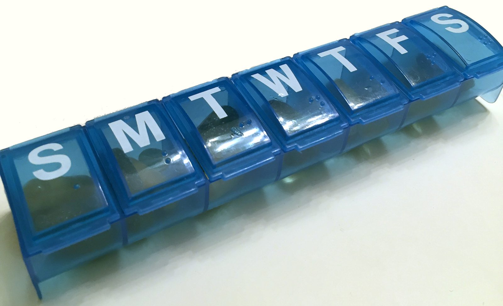 A blue weekly pill organizer with compartments labeled S M T W T F S, photographed at an angle against a plain light background