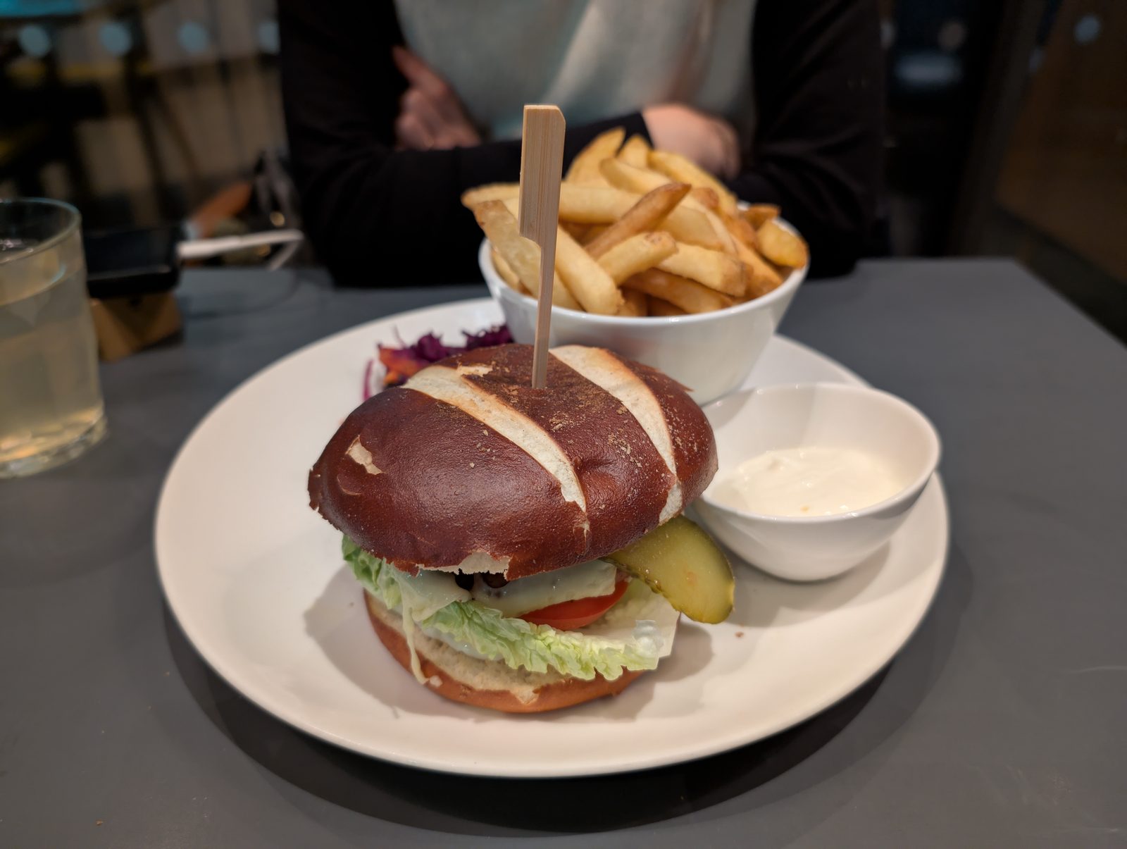 A plated plant based cheeseburger on a pretzel style bun with lettuce, tomato and pickle, served with a bowl of fries and a small dipping sauce