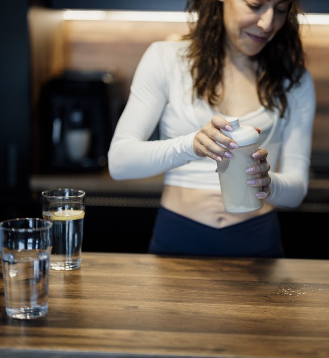 A person in workout clothes stands at a kitchen counter mixing a shake in a shaker bottle, with glasses of water nearby
