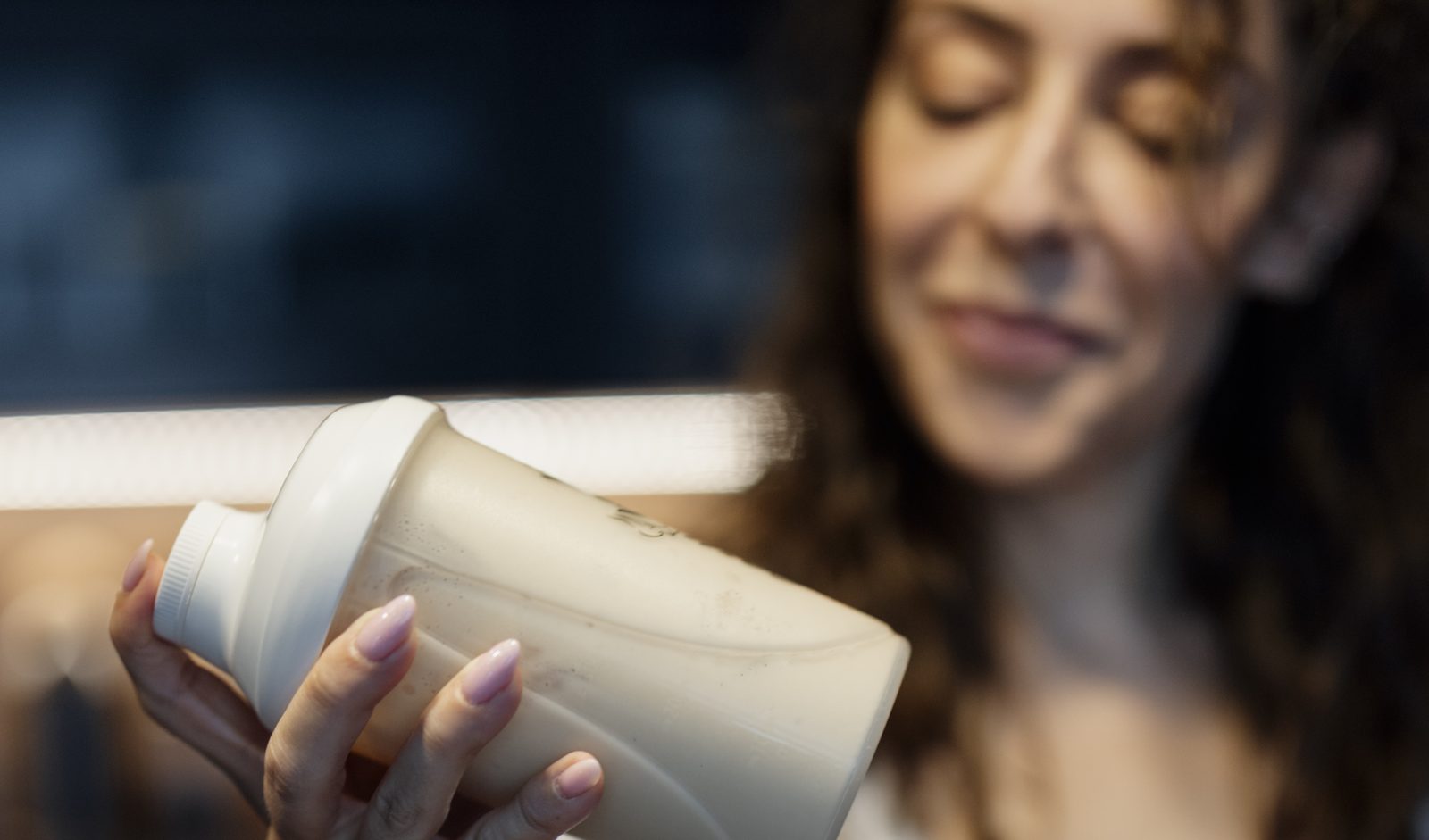 A person holding a protein shaker bottle with a shake mixed inside, a glass with a lemon slice on the counter