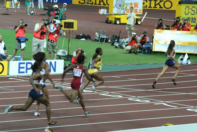 Sprinters lean through the finish line of a 200 meter race at a major outdoor track meet, photographers and officials lining the infield