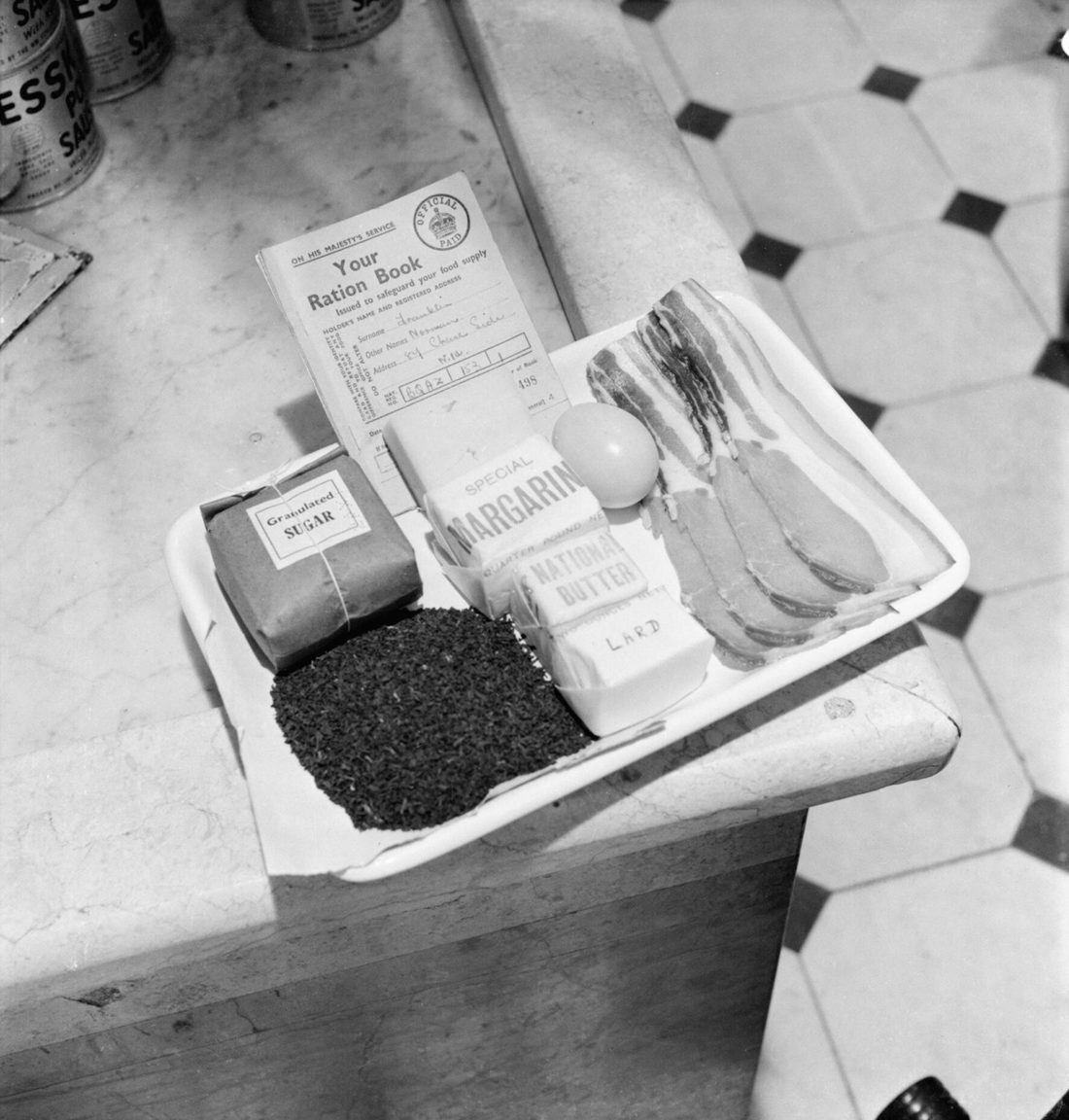A tray holding a British ration book alongside the actual weekly ration of sugar, tea, margarine, butter, lard, eggs, bacon and cheese issued to one adult in 1942