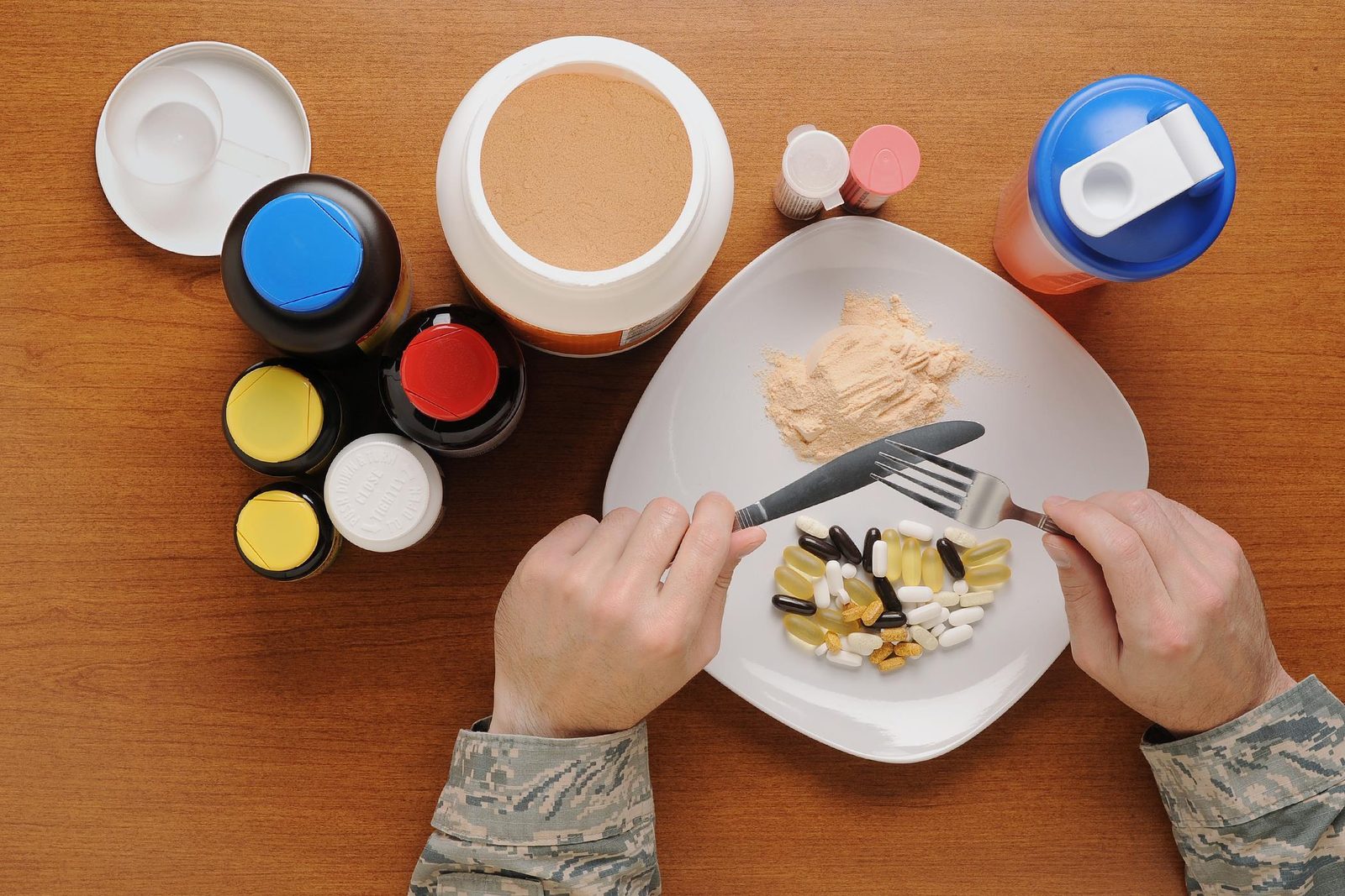 Overhead view of a person in fatigues eating a plate of assorted supplement capsules with a fork and knife, surrounded by protein powder tubs and pill bottles