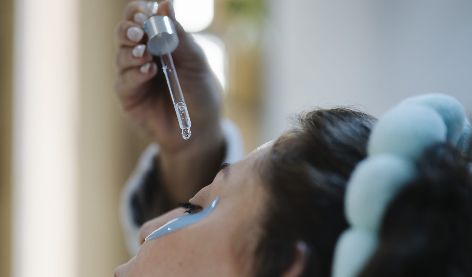 A woman tilts her head back as a drop of serum falls from a glass dropper toward her cheek