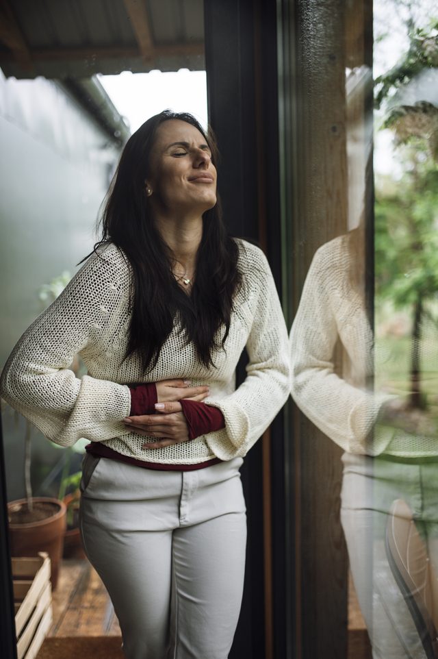 A woman standing by a window holds her stomach with a pained expression